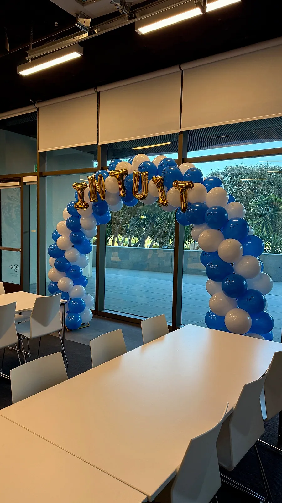 Blue and white balloon arch with gold INTUIT letter balloons framing the window wall of a conference room