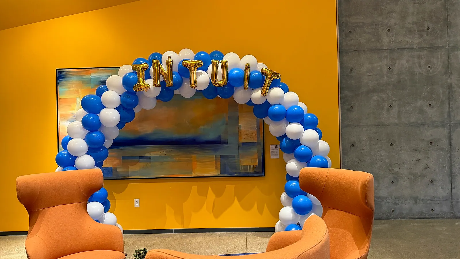 Blue and white balloon arch topped with gold foil letters spelling INTUIT, framing the artwork wall in an Intuit lobby with orange armchairs