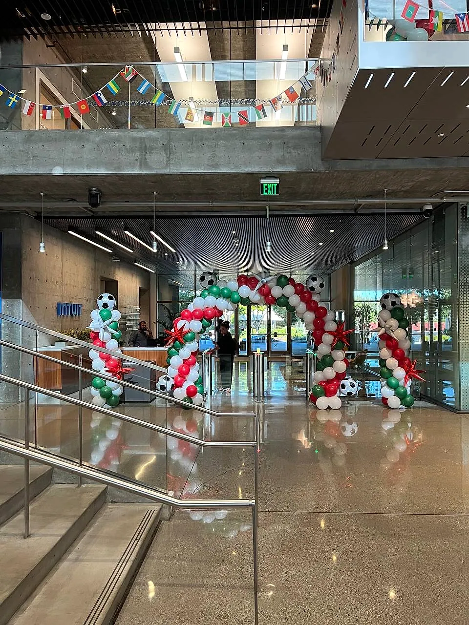 Red, green and white balloon arch with soccer-ball foil balloons at a glass building entrance, under a run of national flag bunting