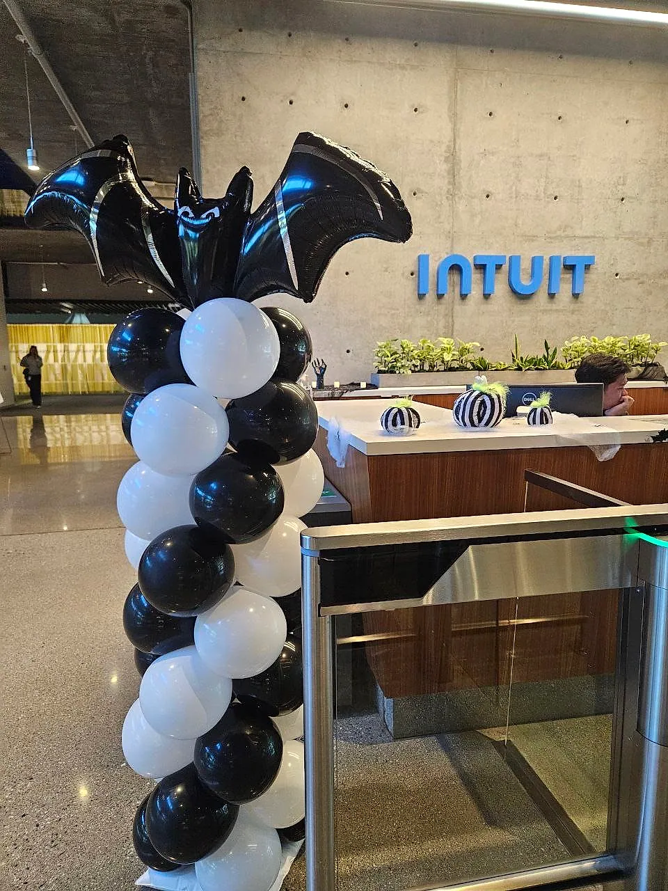 Black and white balloon column topped with a large bat foil balloon, standing at a reception desk beneath the blue INTUIT lobby sign
