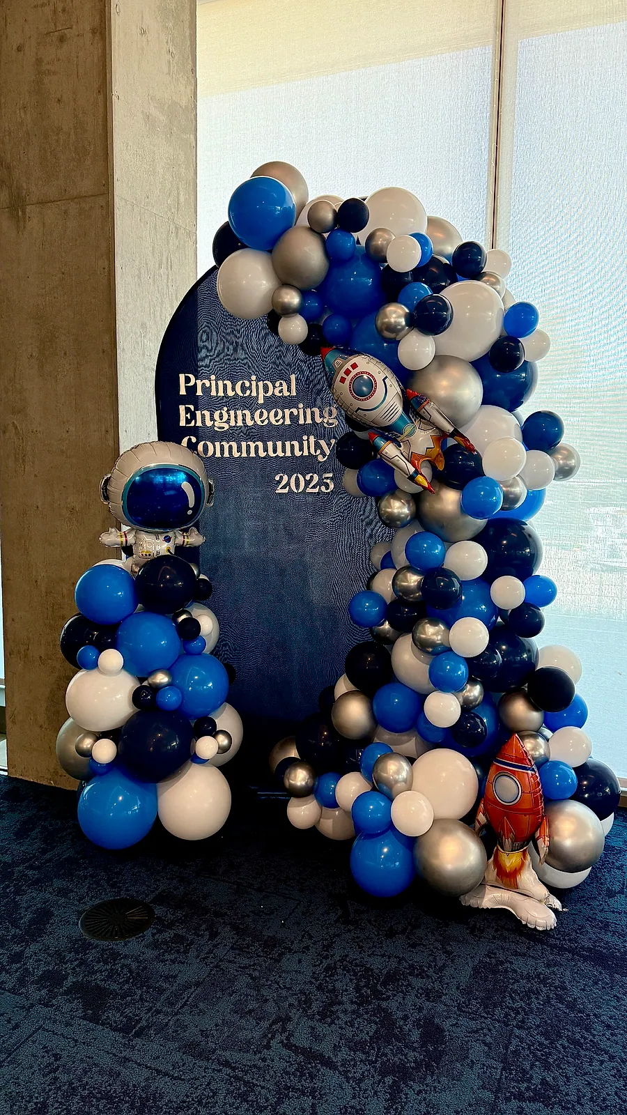 Navy, blue, silver and black organic balloon garland wrapping an oval Principal Engineering Community 2023 sign, with rocket and planet foil balloons