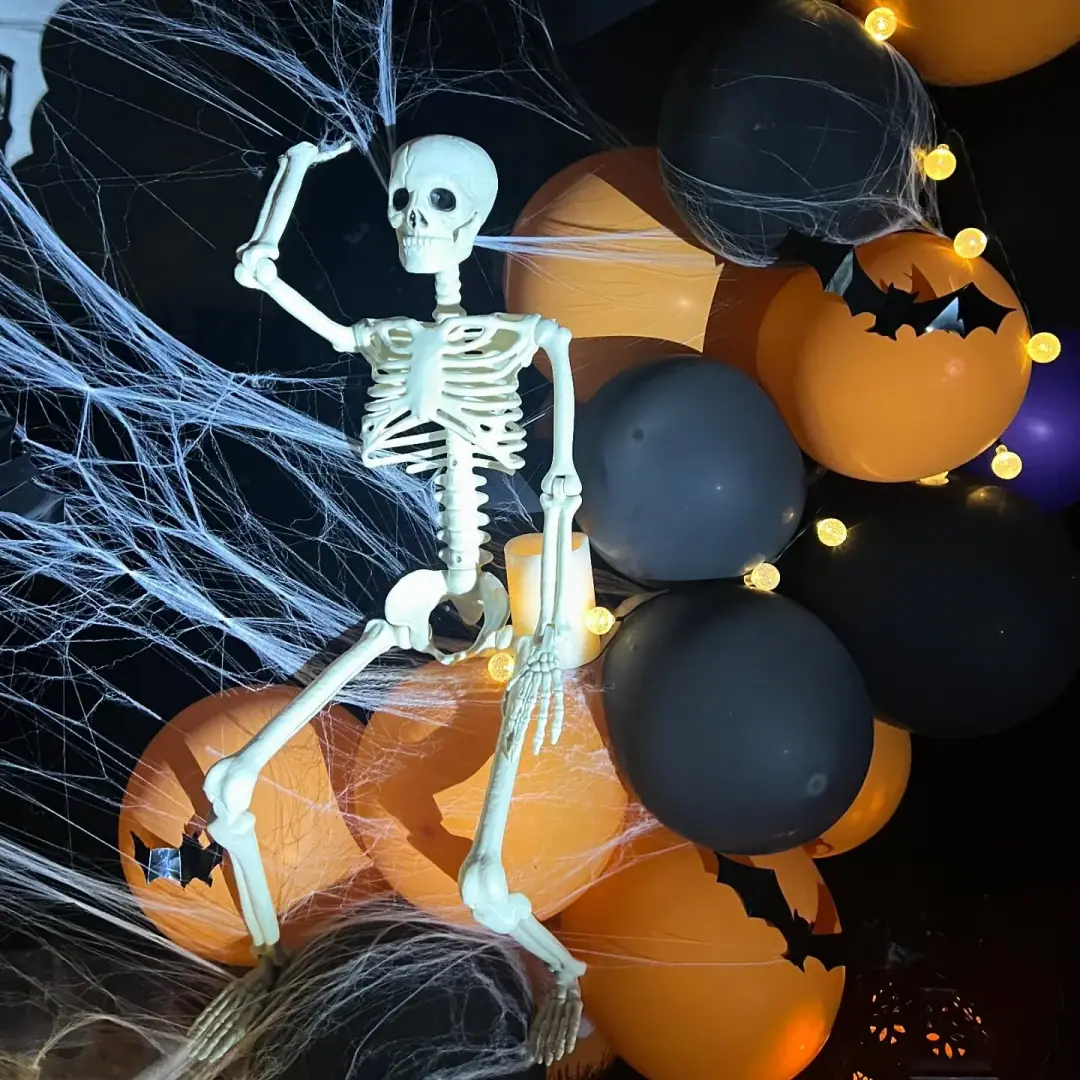 Close-up of black and orange Halloween balloons with bat cutouts, stretched cobweb, fairy lights and a posed skeleton