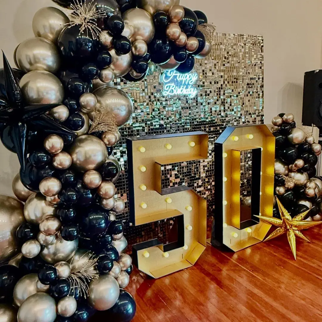 Black, chrome gold and silver balloons in mixed sizes beside light-up gold number 50 in front of a sequin wall with a neon sign