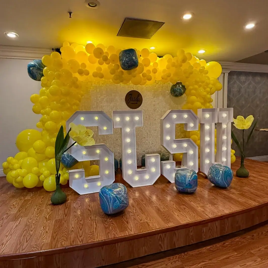 Yellow balloon wall covering the back of a stage behind light-up marquee letters and blue marbled balloons