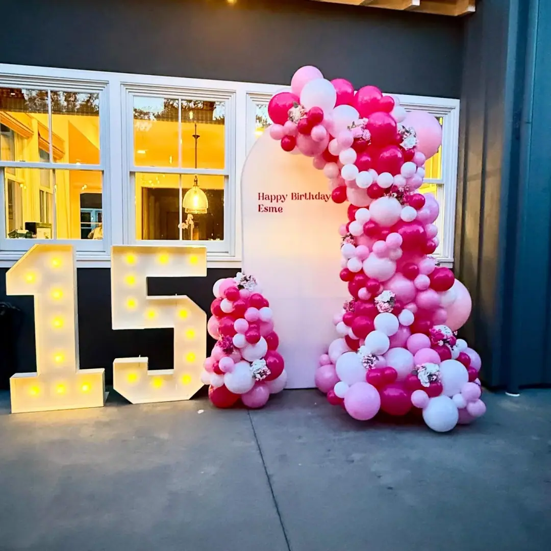 Light-up marquee number 15 beside a pink, fuchsia and white balloon garland and a birthday sign panel