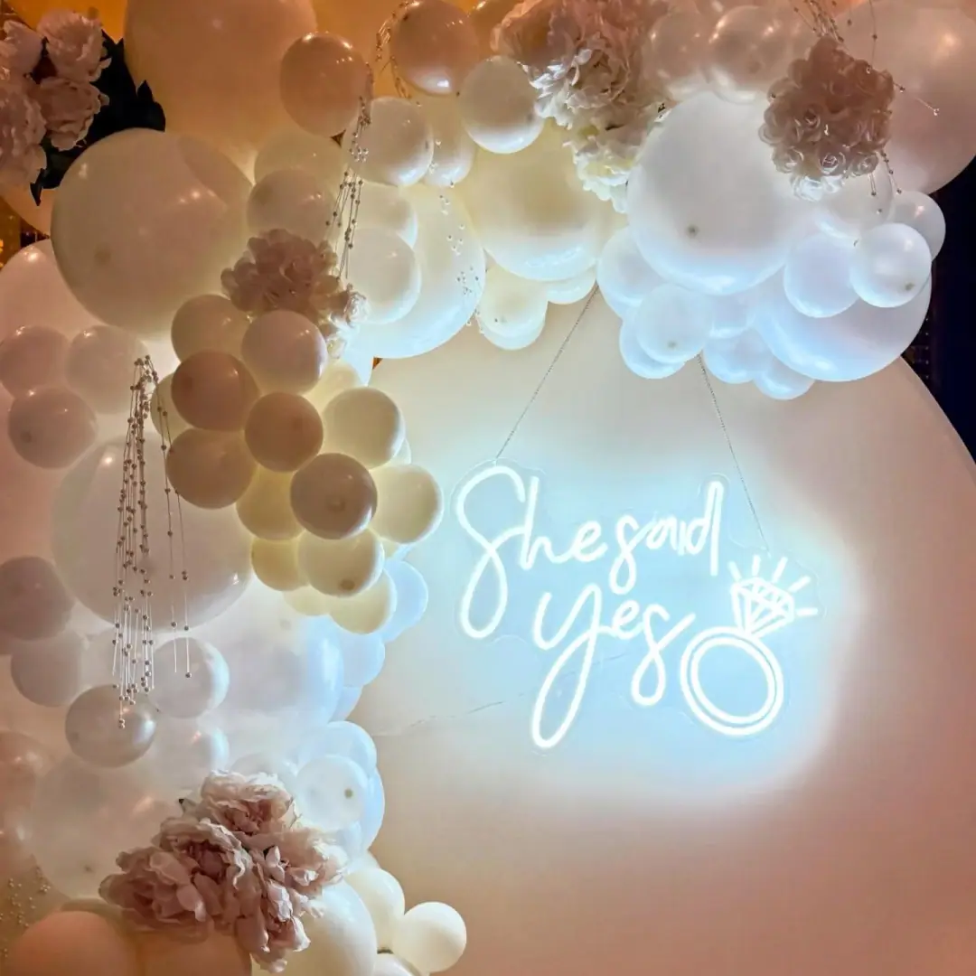 Close detail of a layered balloon installation with fresh blooms and a glowing neon sign