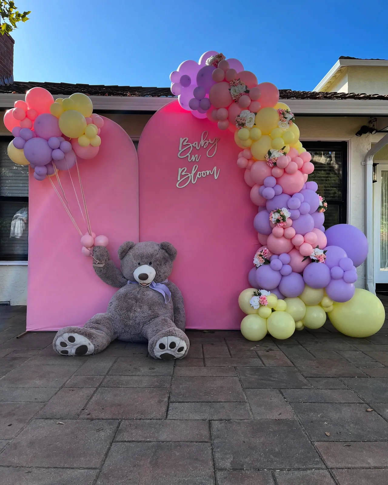 Pink, lilac and yellow baby shower balloon garland on a Baby in Bloom backdrop with a giant teddy bear