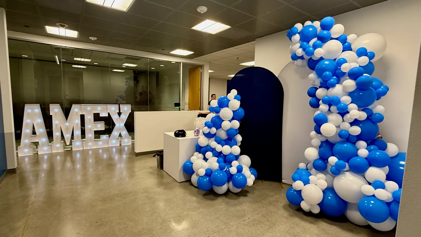 Light-up AMEX marquee letters beside a blue and white balloon garland on a navy arch at an office reception
