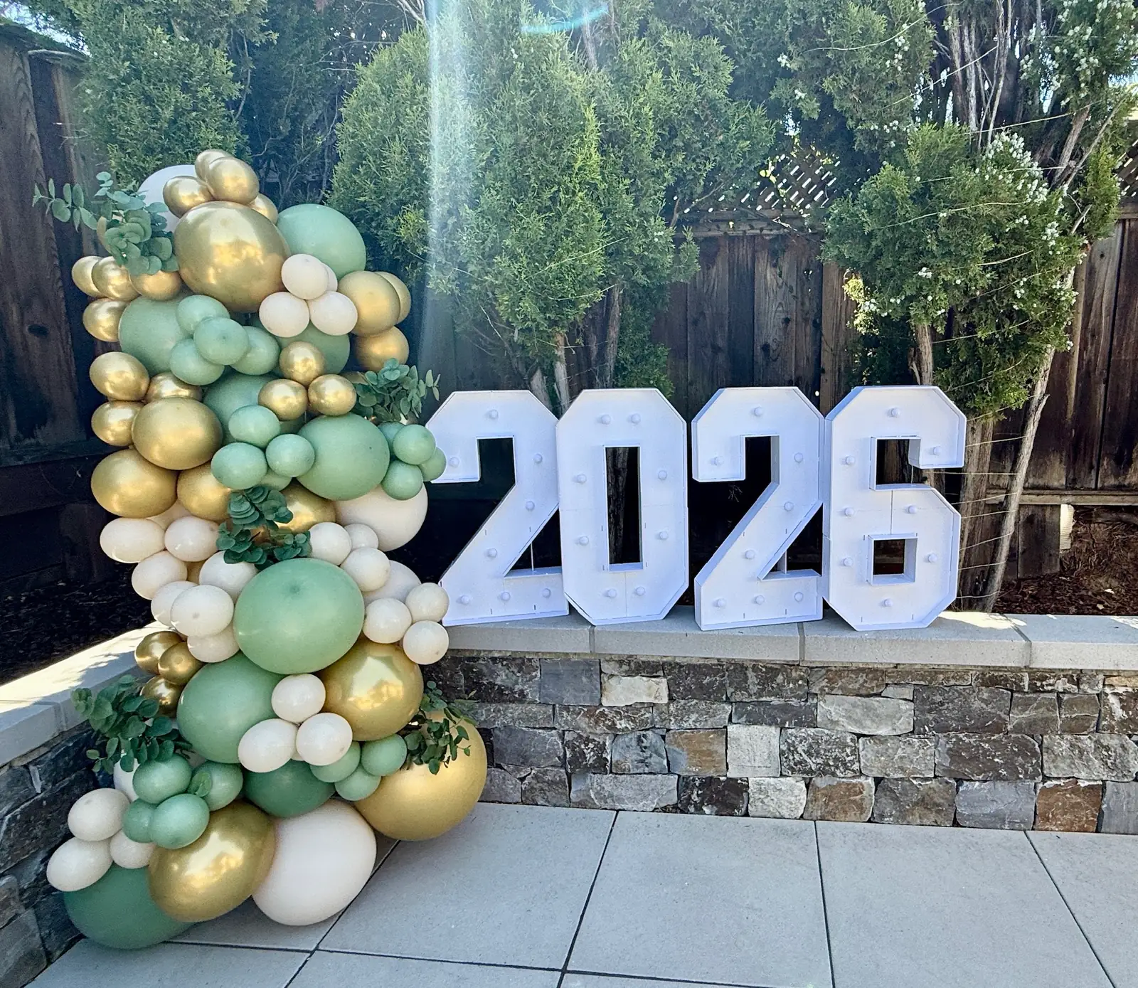 Sage, white and chrome gold balloon garland beside light-up 2026 marquee numbers on a patio