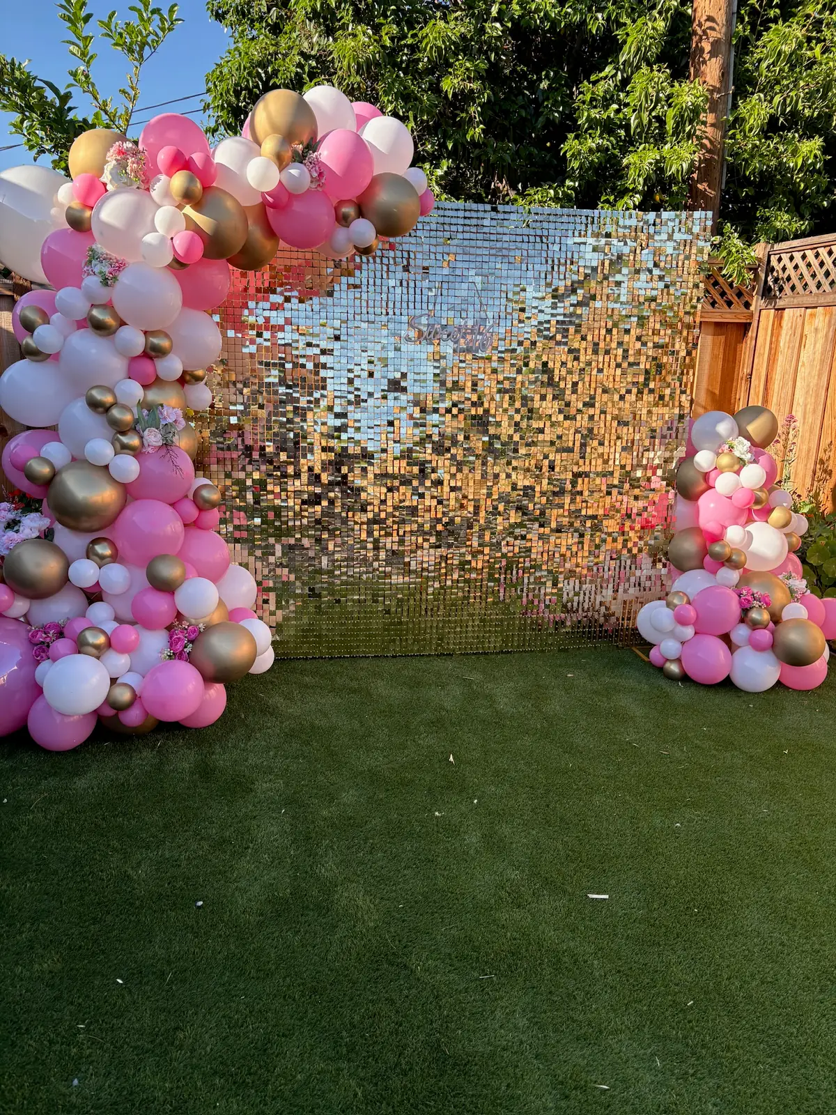 Pink, blush and gold balloon garland beside a silver sequin wall on a backyard lawn