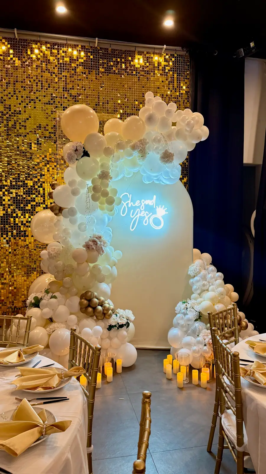 White, ivory and champagne balloon garland on an ivory arch with a She Said Yes neon sign in front of a gold sequin wall